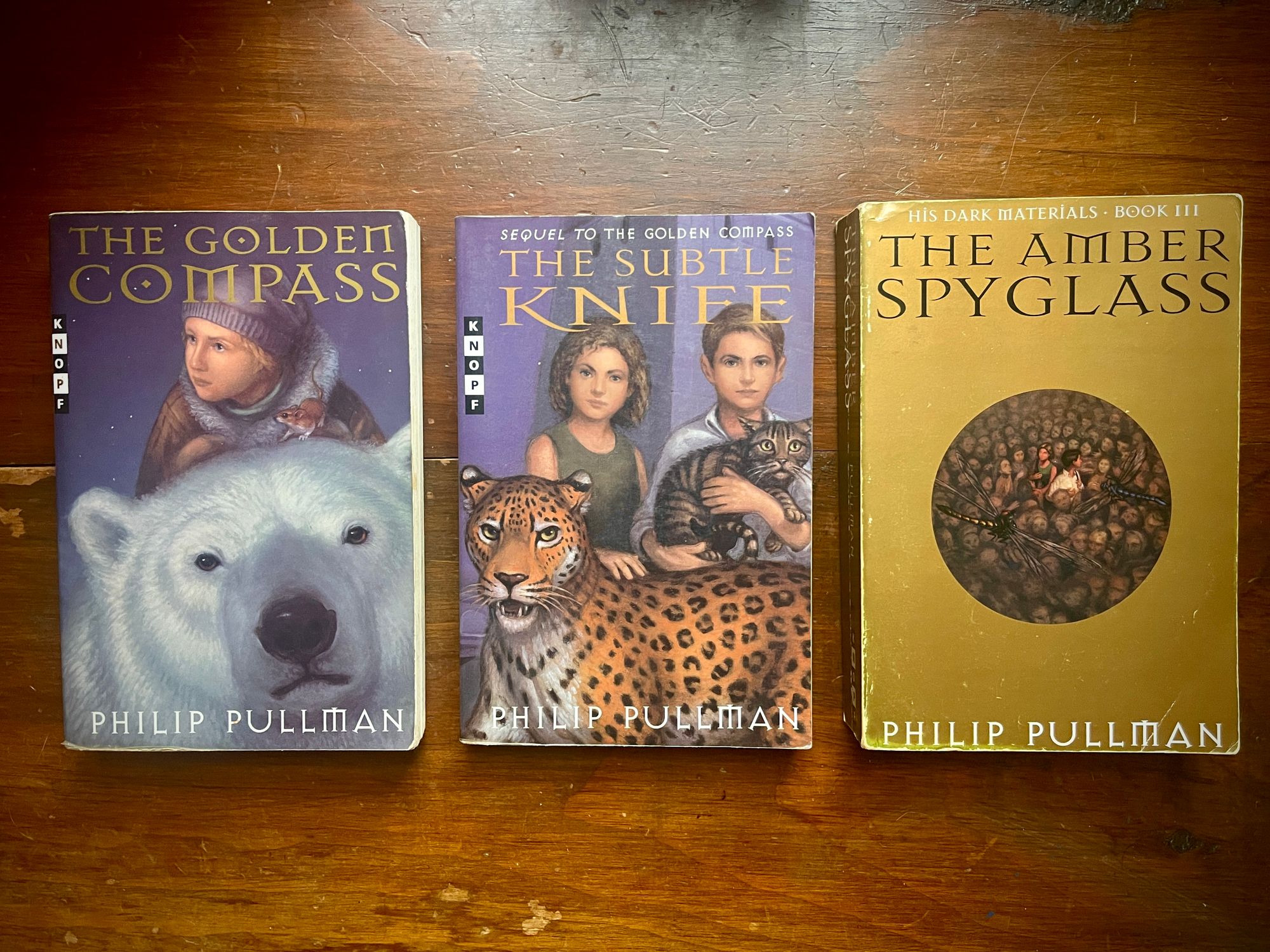 Three novels on a table: The Golden Compass, The Subtle Knife, and The Amber Spyglass.