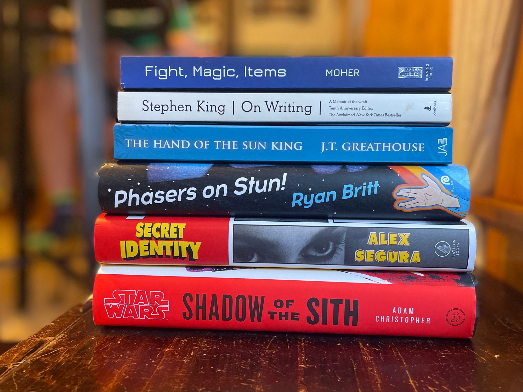 A stack of blue and red books: Fight Magic, Items, Stephen King’s On Writing, J.T. Greathouse’s The Hand of the Sun King, Phasers on Stun by Ryan Britt, Secret Identity by Alex Segura, and Shadow of the Sith by Adam Christopher